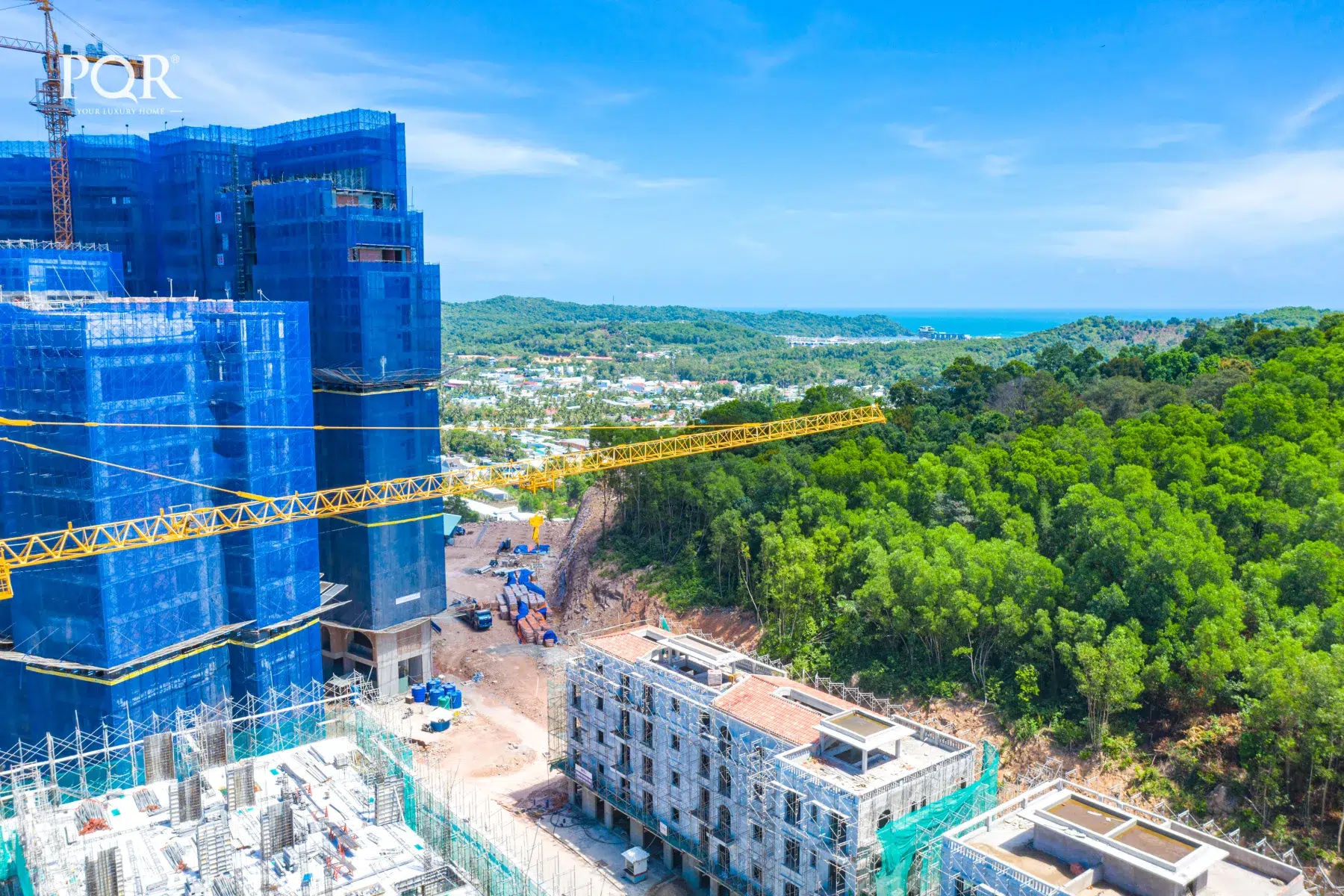 Ảnh giai đoạn xây dựng Hillside với công trình bọc lưới xanh bên trái, sườn đồi bên phải và biển ở xa Địa thế và hướng căn Hillside: đọc từ tổng thể đến từng căn